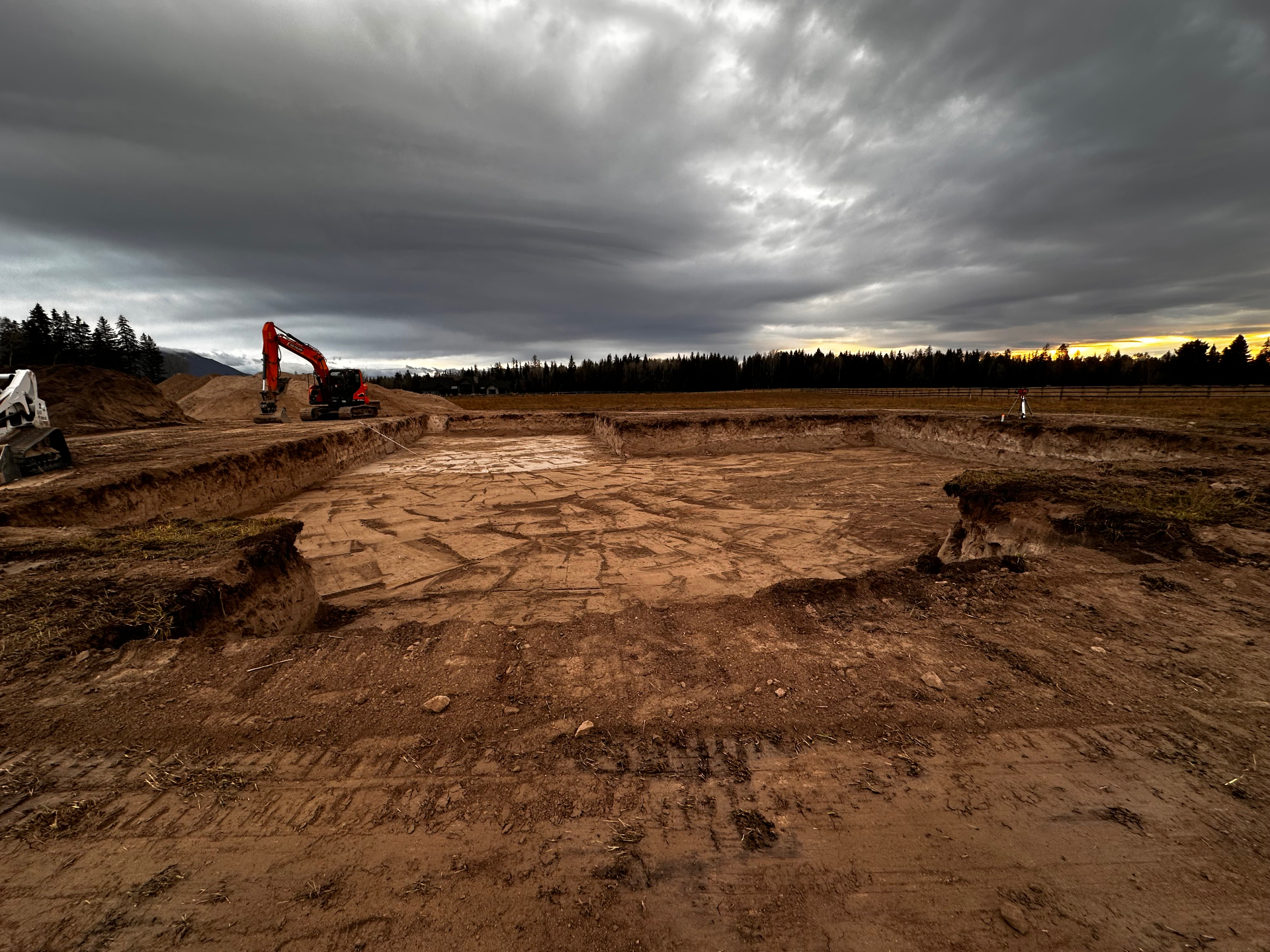 Excavated foundation pad at dusk with excavator and mountains in background